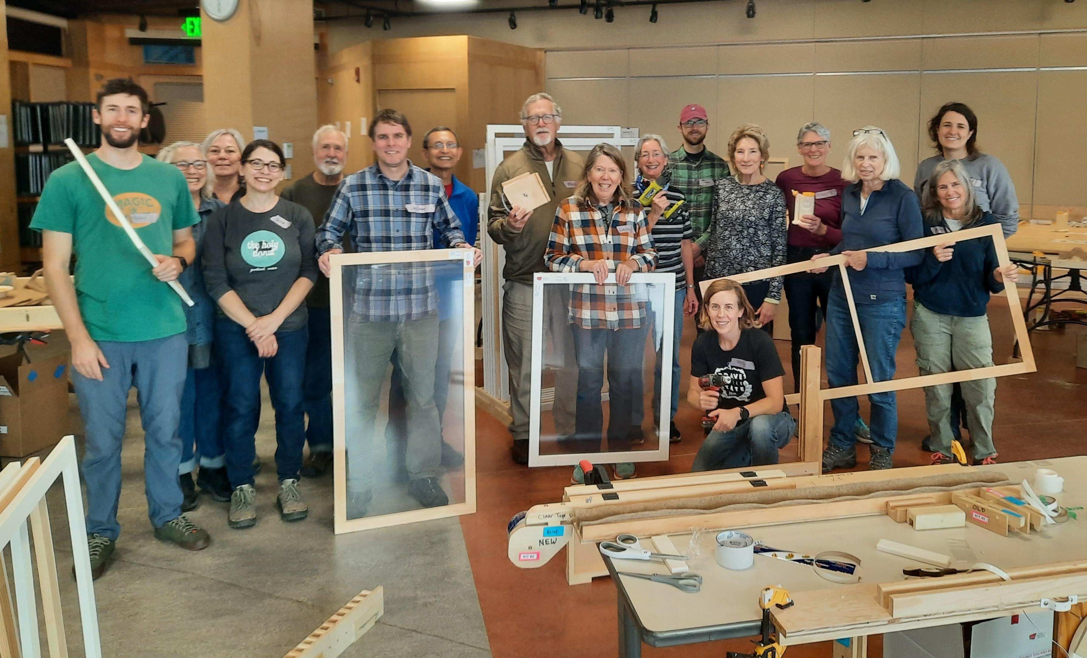 17 people pose for a group photo with wooden window frames, some covered with plastic, and other insert parts. There's a table in the foreground with project materials like tape and scissors, and custom wooden jigs to aid alignment. The space is ECHO's Lakeside room.